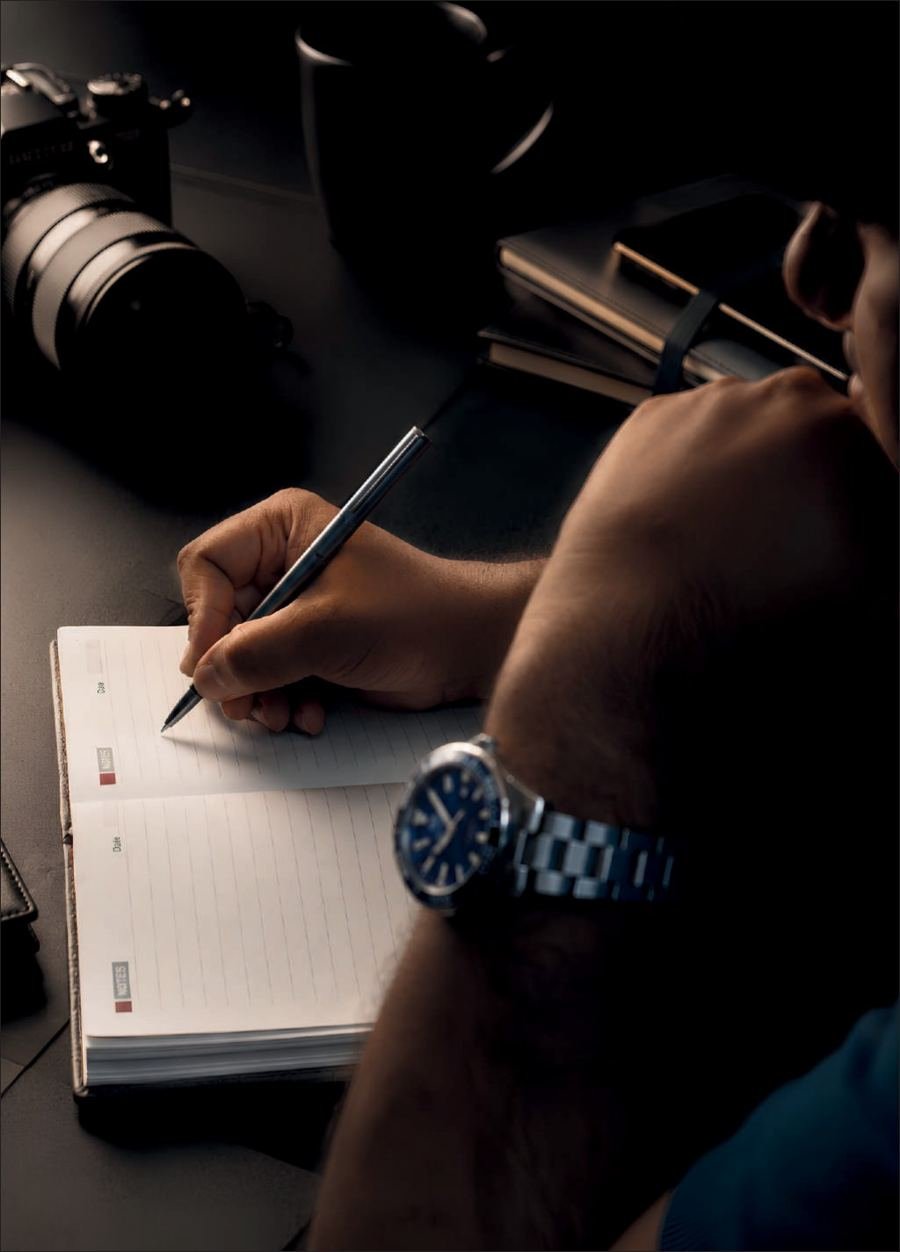 A hand writing in a notebook with a silver pen beside a camera